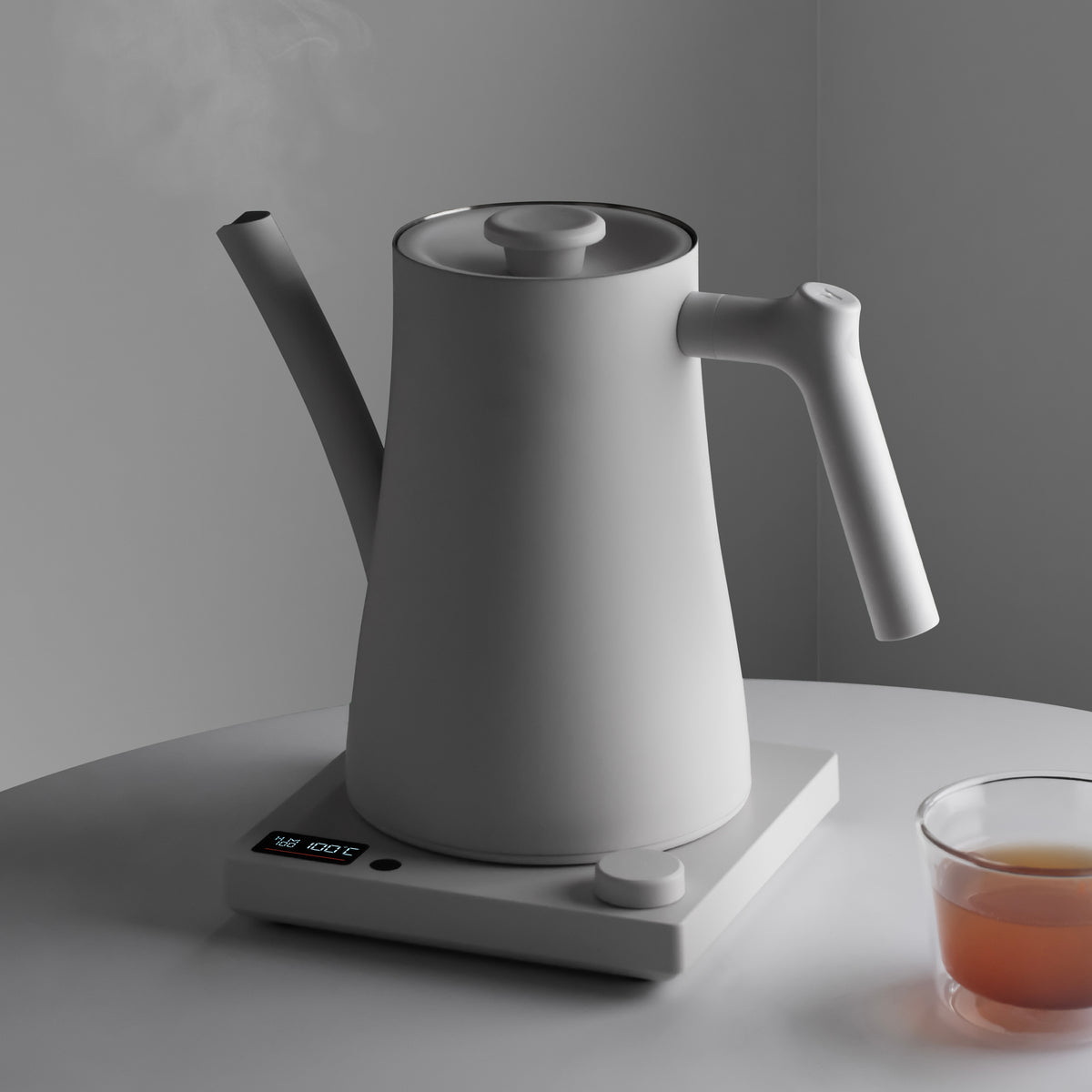 White AURA 1.5 L kettle on its base, with steam rising beside a glass of tea